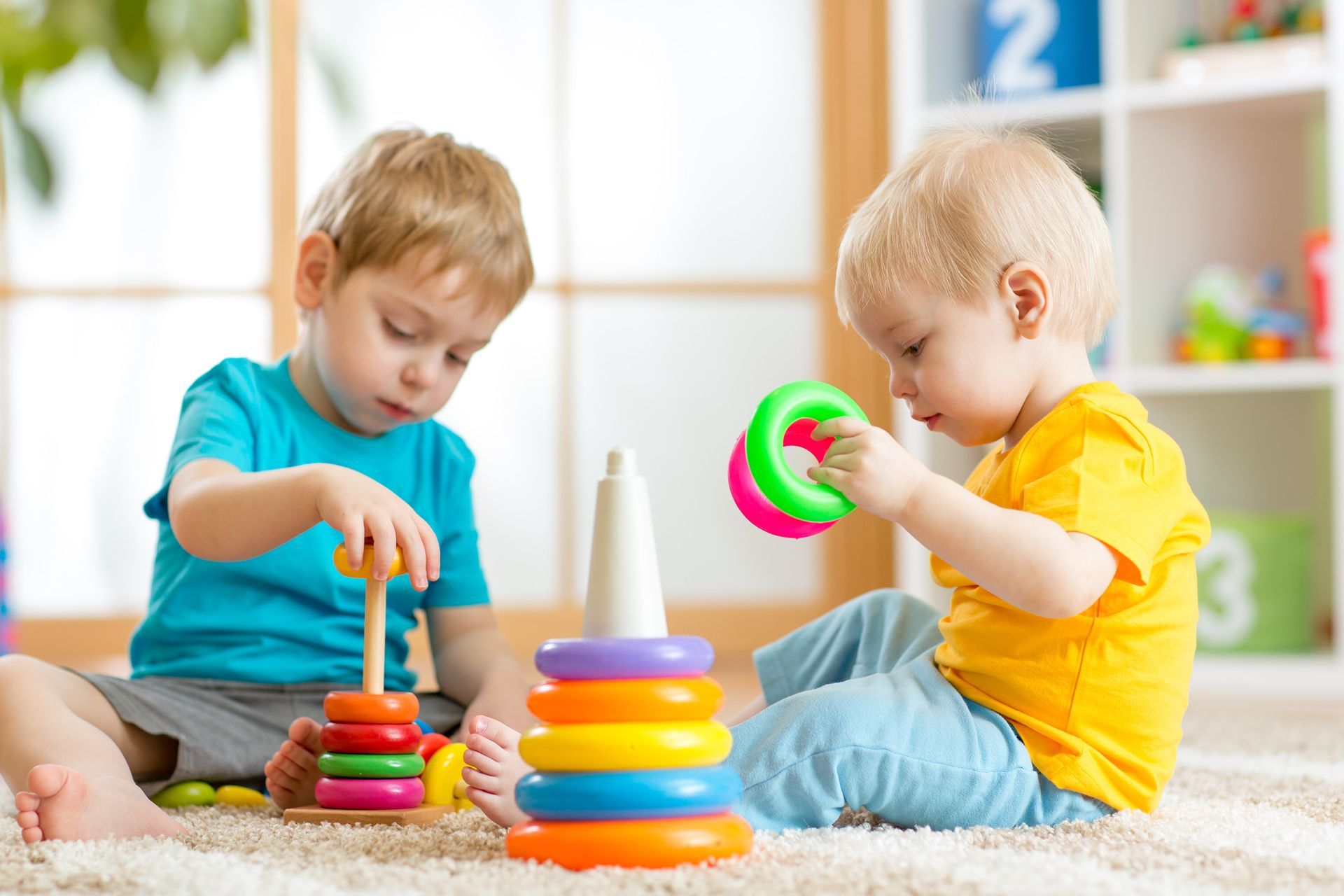 Two toddlers sitting on a rug playing with colorful stacking rings in a well-lit room.