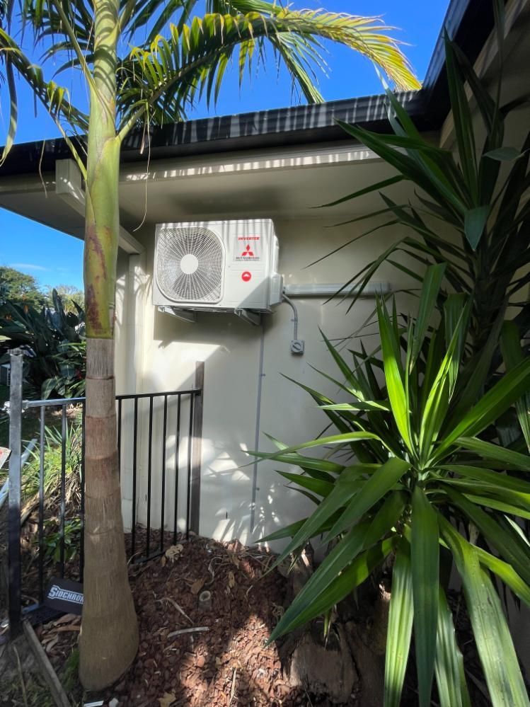 A White Air Conditioner is Mounted on the Side of a House Next to a Palm Tree — Condie Refrigeration & Air Conditioning In Pottsville, NSW