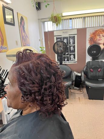 Hair salon interior with a client in a black cape and curled burgundy hair, styling chairs and posters in the background
