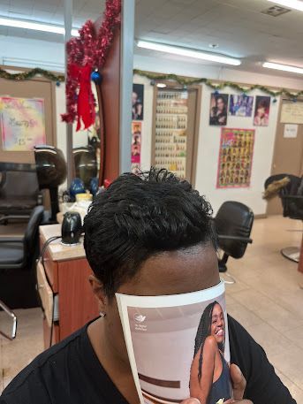 Person in a salon chair with short black hair, looking down at a magazine in a brightly lit salon.