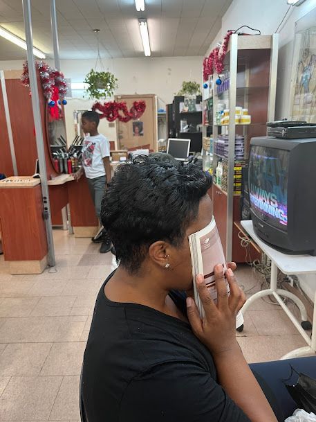 Person seated in a salon waiting area, holding a book, with a TV and reception desk nearby.