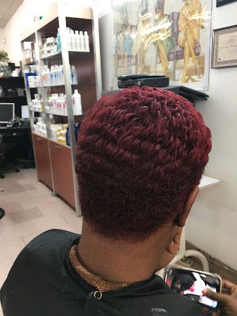 Person with short burgundy waves seen from behind in a salon, with product shelves and posters in the background