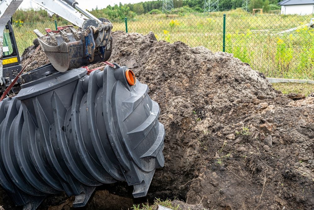 Excavator burying a large gray septic tank in dirt, near a fence.