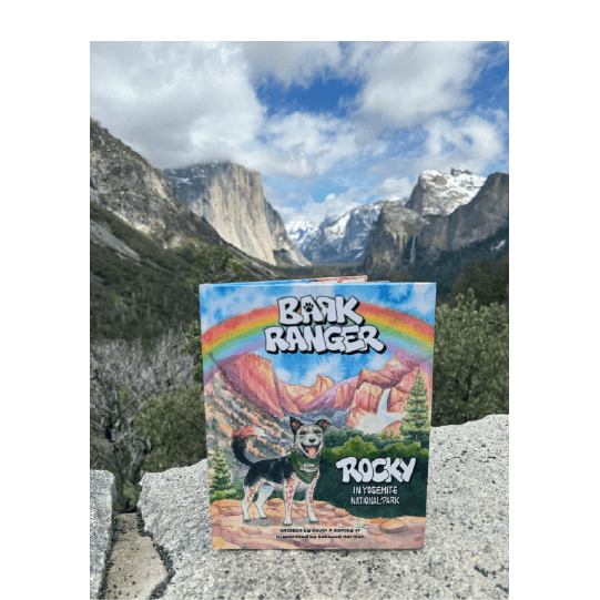 Book standing up on a rock ledge with mountains in the background; the book cover shows a dog with the same mountains and a rainbow. Text: Bark Ranger. Rocky in Yosemite National Park.