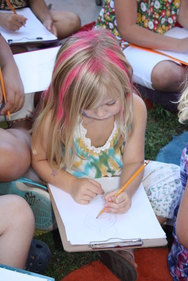 Children sitting together on the grass and drawing. Text: Learning with illustrators