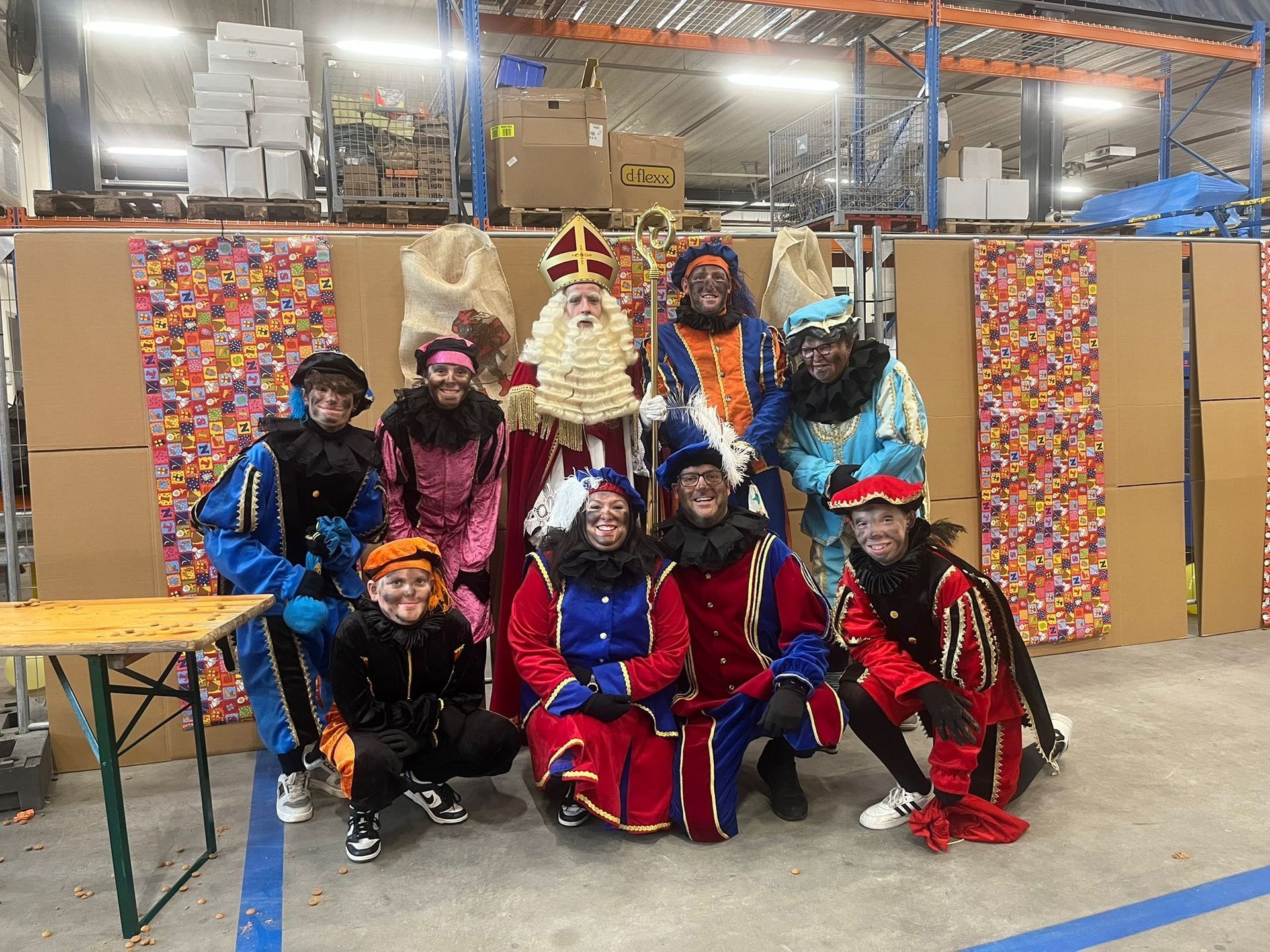 Een groep in kleurrijke kostuums poseert binnen tijdens een feestelijk evenement met een versierd podium als achtergrond.