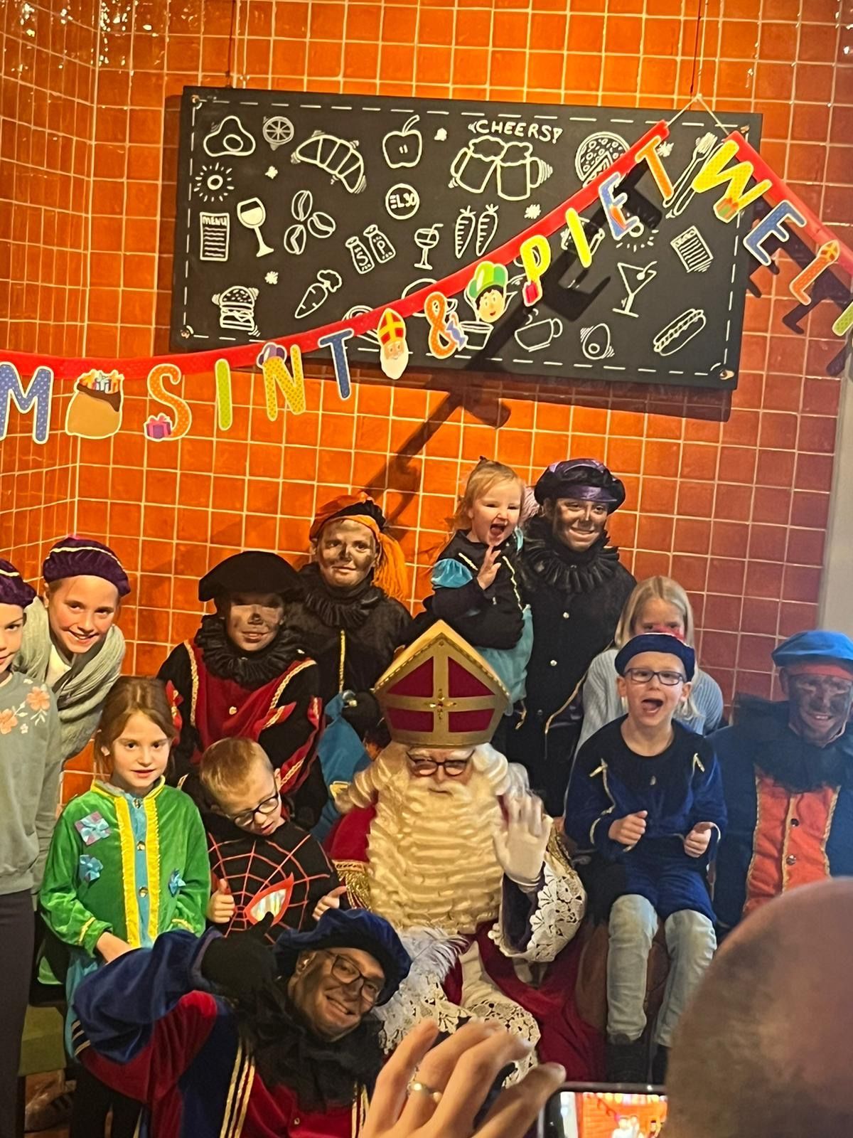 Kinderen poseren met de sint voor een schoolbord en een oranje bakstenen muur, met daarboven een feestelijke banner.