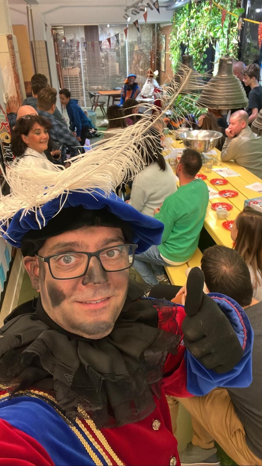 Een persoon in een kleurrijk kostuum maakt een selfie op een drukbezocht feestelijk binnenfeest met versierde tafels.