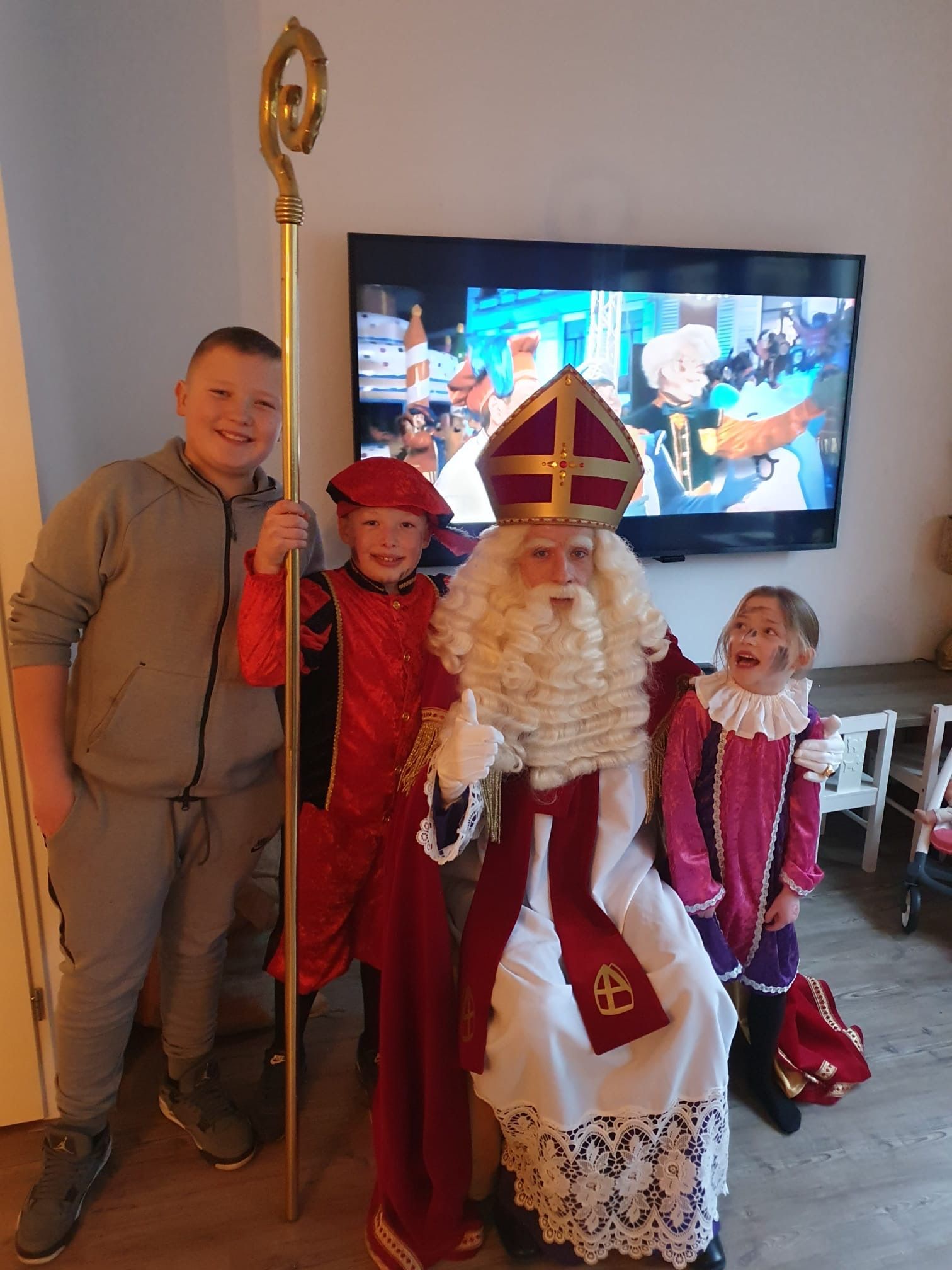 Drie mensen in feestelijke kostuums met Sinterklaas, staand binnen voor een tv.