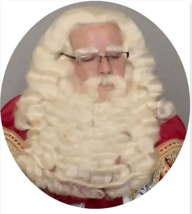 Persoon in een kerstmanpak met witte krullende baard en bril, gekleed in rood en goud.