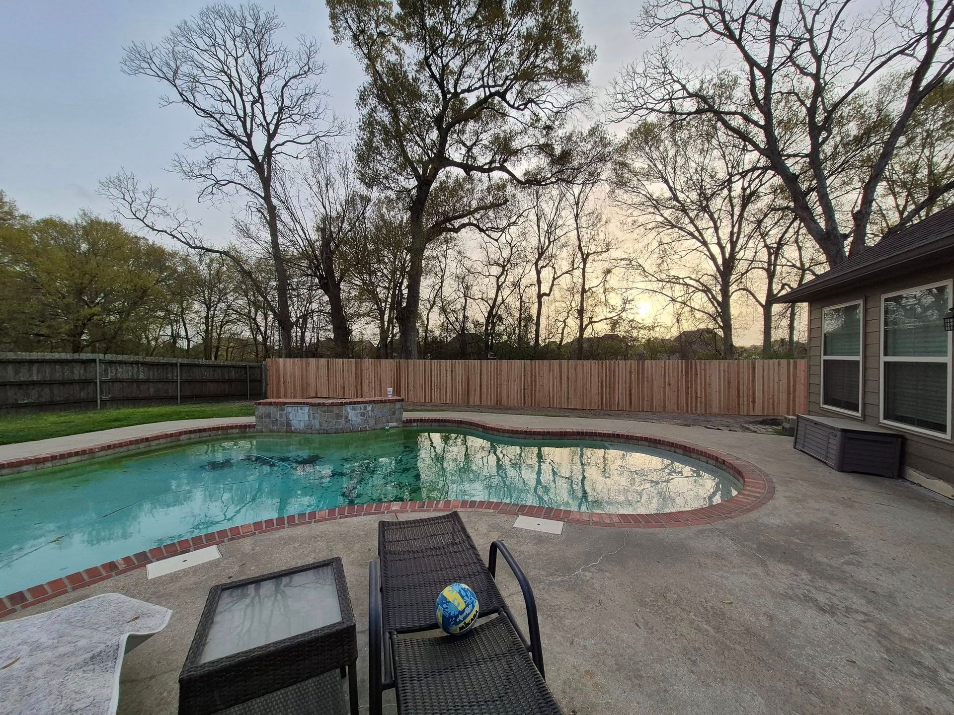 Pool and patio area with a wooden fence. Trees and a sunset in the background.