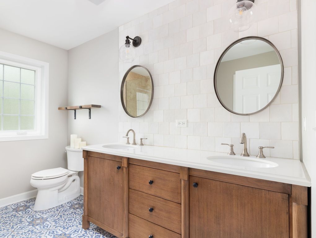 Bathroom with wooden vanity, white tiled wall, two round mirrors, and blue patterned floor tiles.