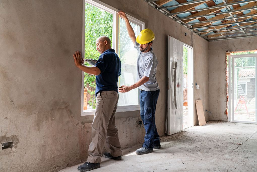 Two people installing a window in a room under construction; one measures, one steadies the frame.