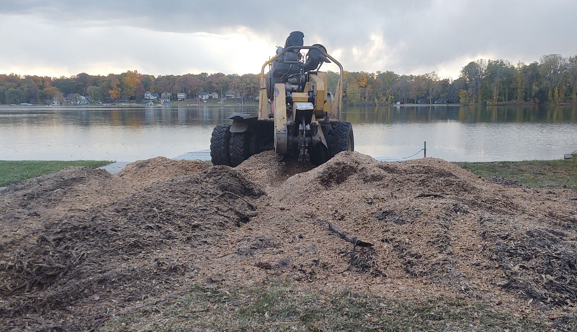 A tree stump grinder is grinding wood into wood chips on the edge of a lake.