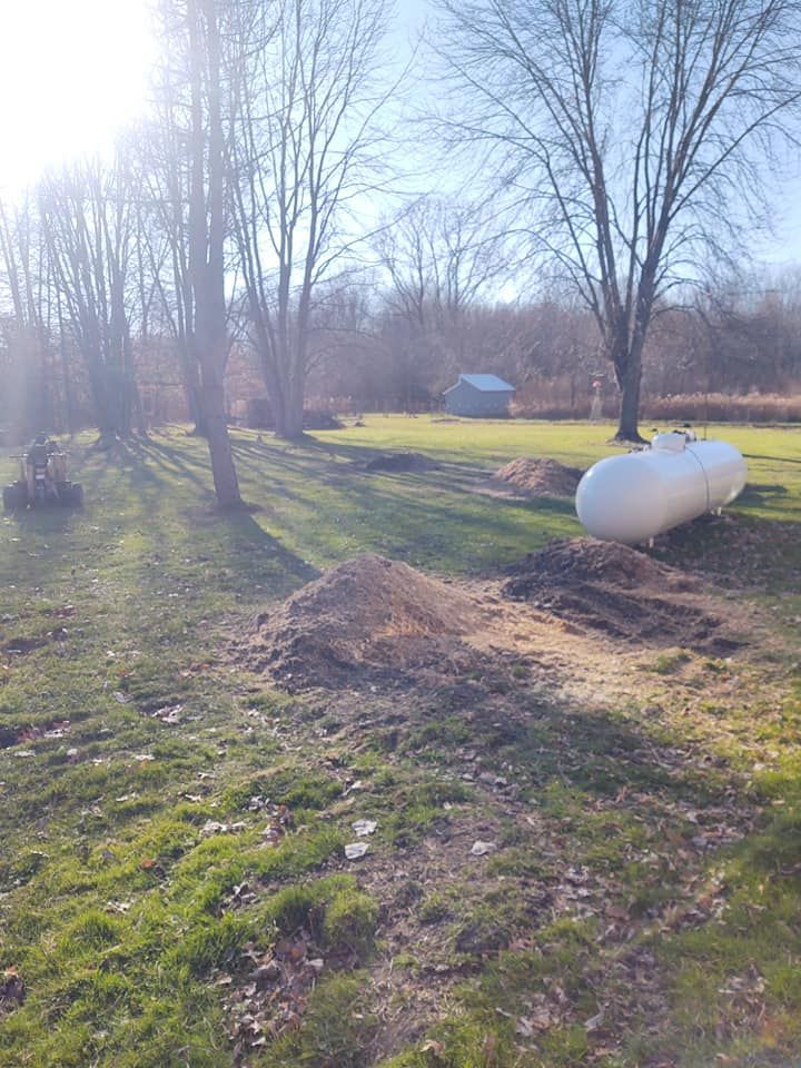 A large white propane tank in a grassy yard, dirt piles, bare trees, and a small blue shed.
