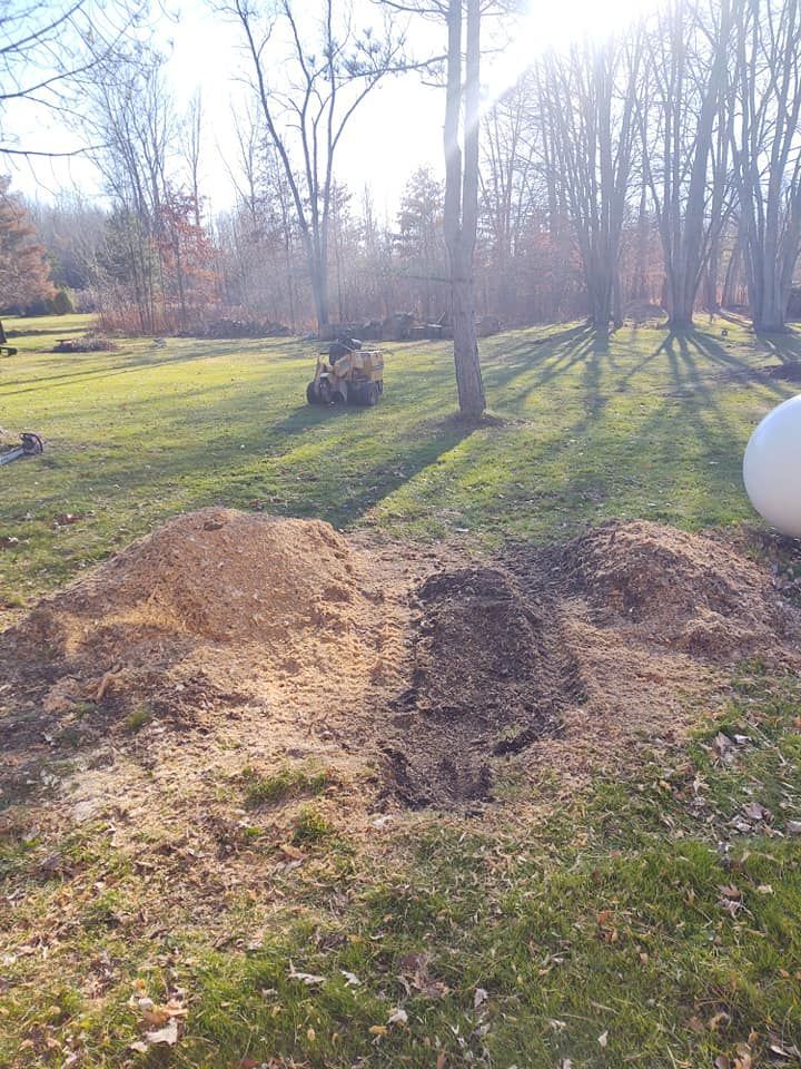 Piles of dirt and mulch in a sunny yard with a lawnmower in the distance, trees, and a propane tank.