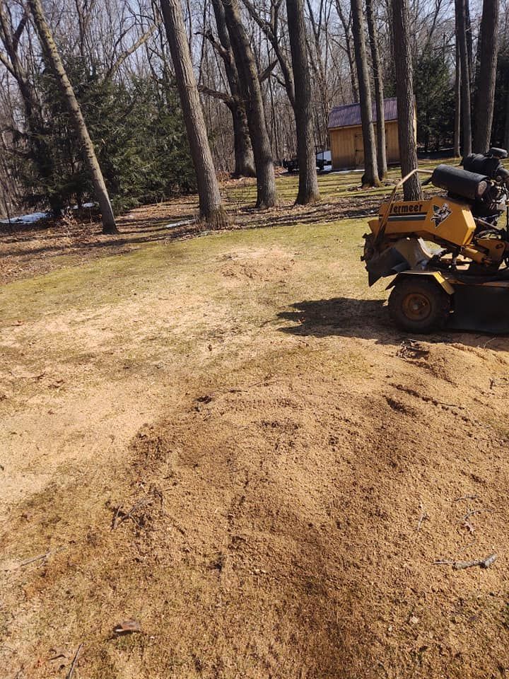 A yellow stump grinder sits next to a pile of wood chips in a grassy yard, trees in the background.
