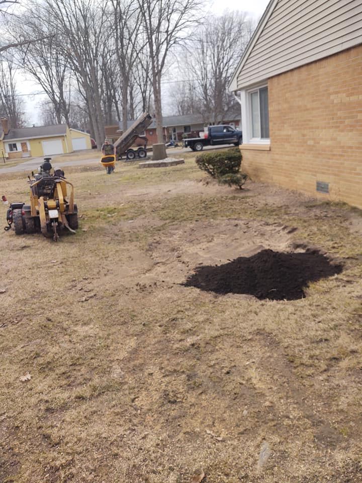 A large hole in a lawn, next to a house. A small excavator and dump truck are visible in the background.