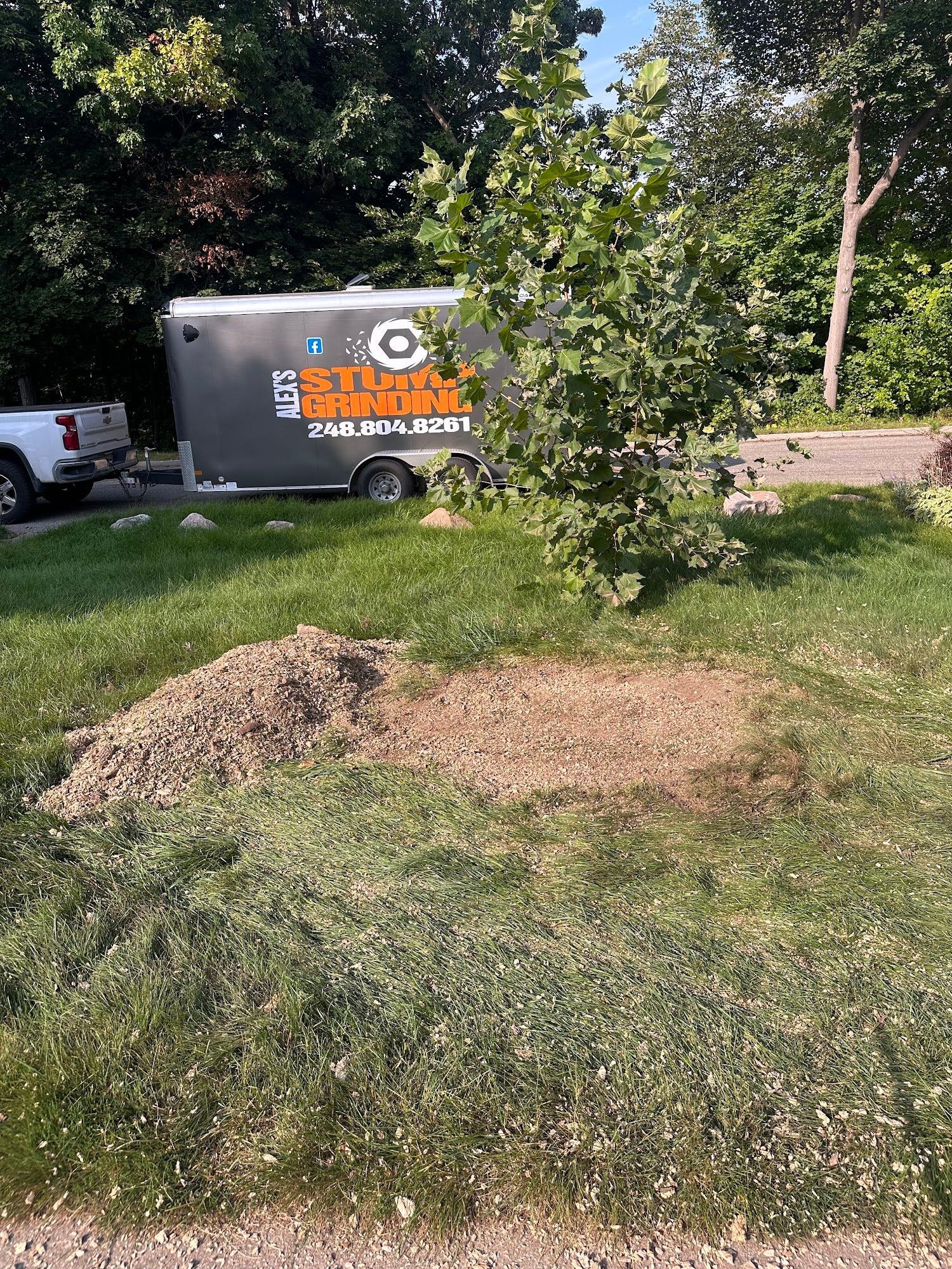 Pile of dirt and bare earth spot in grass with tree, trailer, and truck in the background.