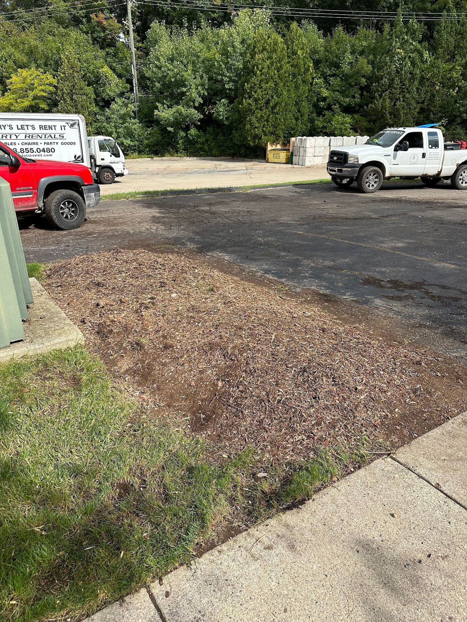 Gravel area next to a sidewalk with trucks and buildings in the background. Green grass on the left.