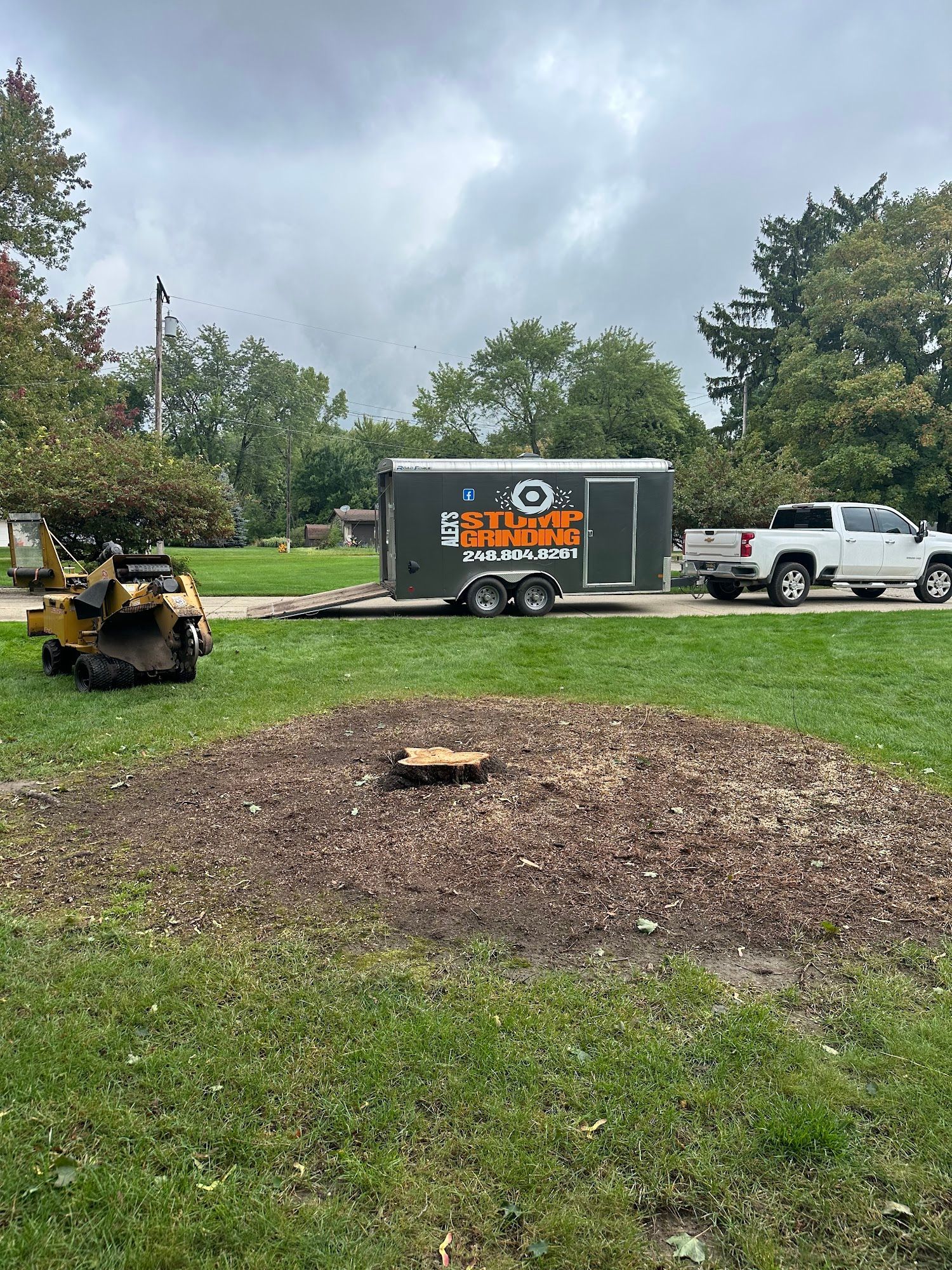 Tree stump after removal, with a stump grinder, trailer, and truck on a grassy lawn. Overcast sky.