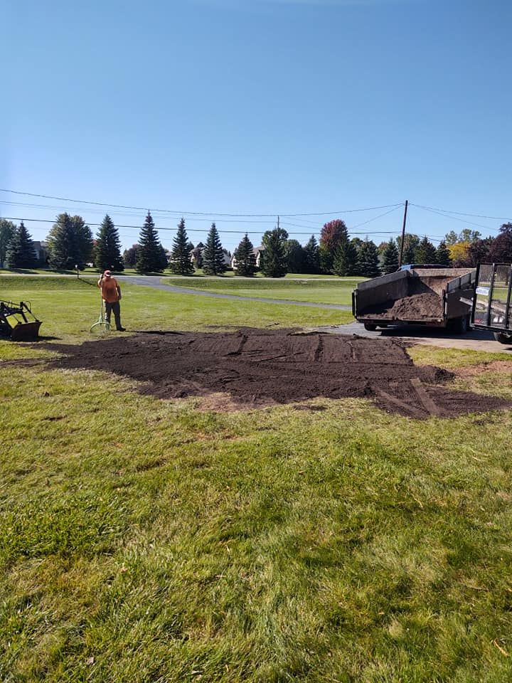 A person spreads dark soil on a lawn near a trailer, under a blue sky.