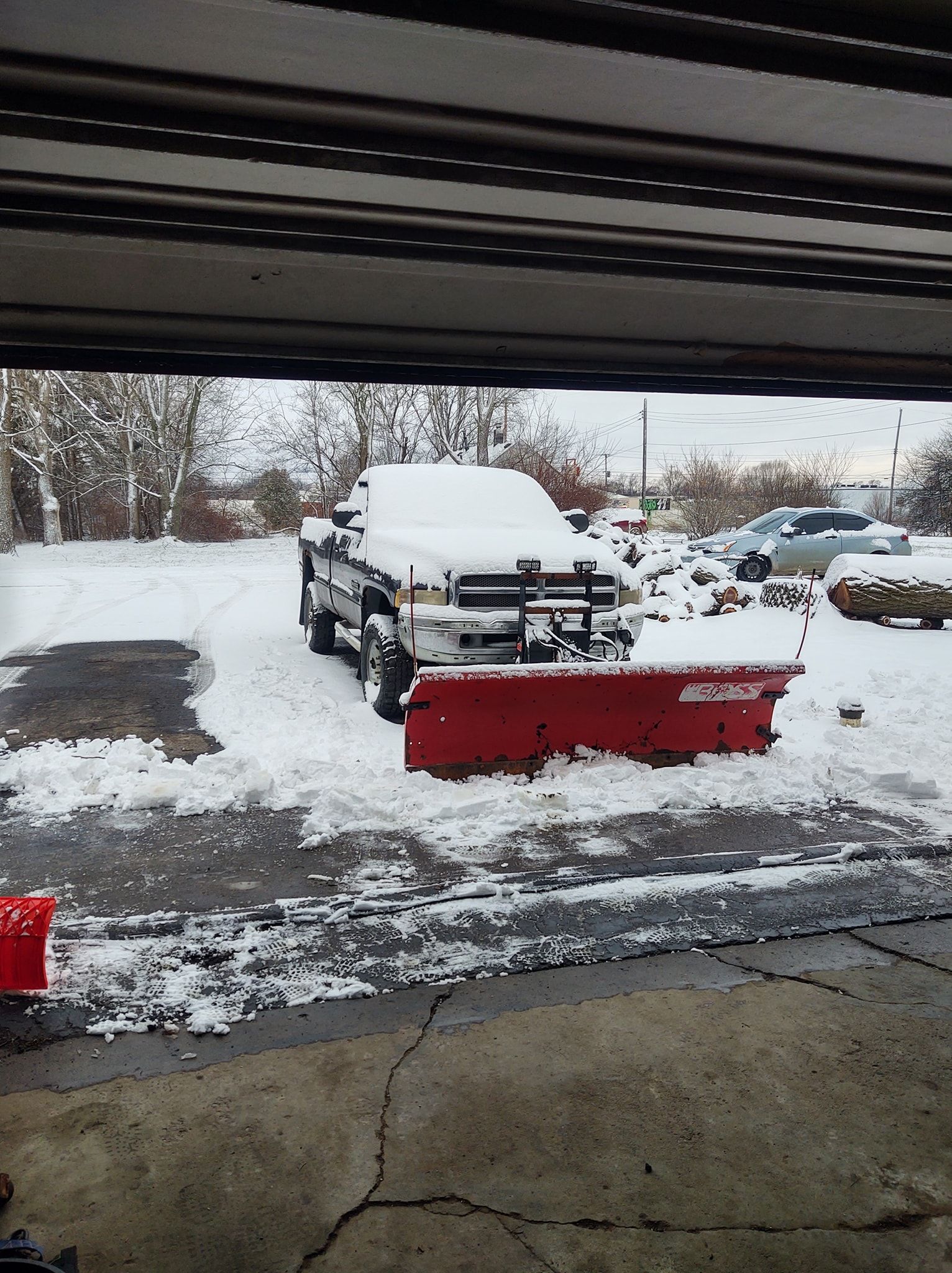 Snow-covered truck with a red snowplow parked in a driveway. Snow is on the ground.