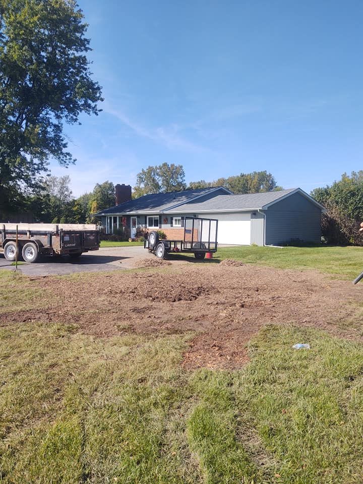 House with trailers on driveway, recently tilled dirt in front. Green grass, blue sky.