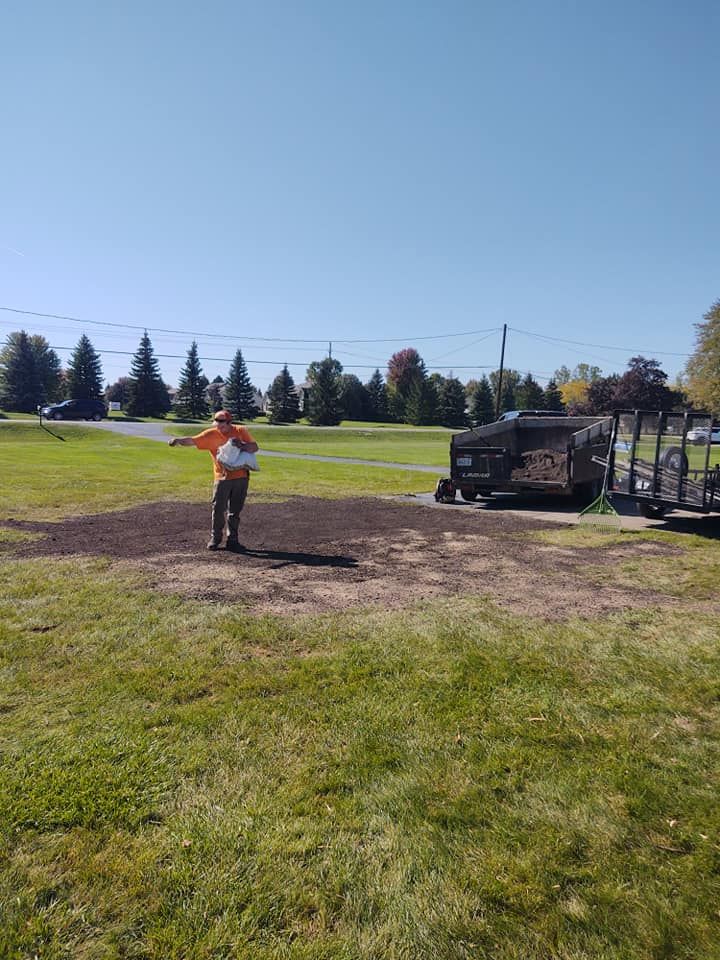 Person spreading material on dark soil patch in a grassy field. Equipment is nearby on a sunny day.