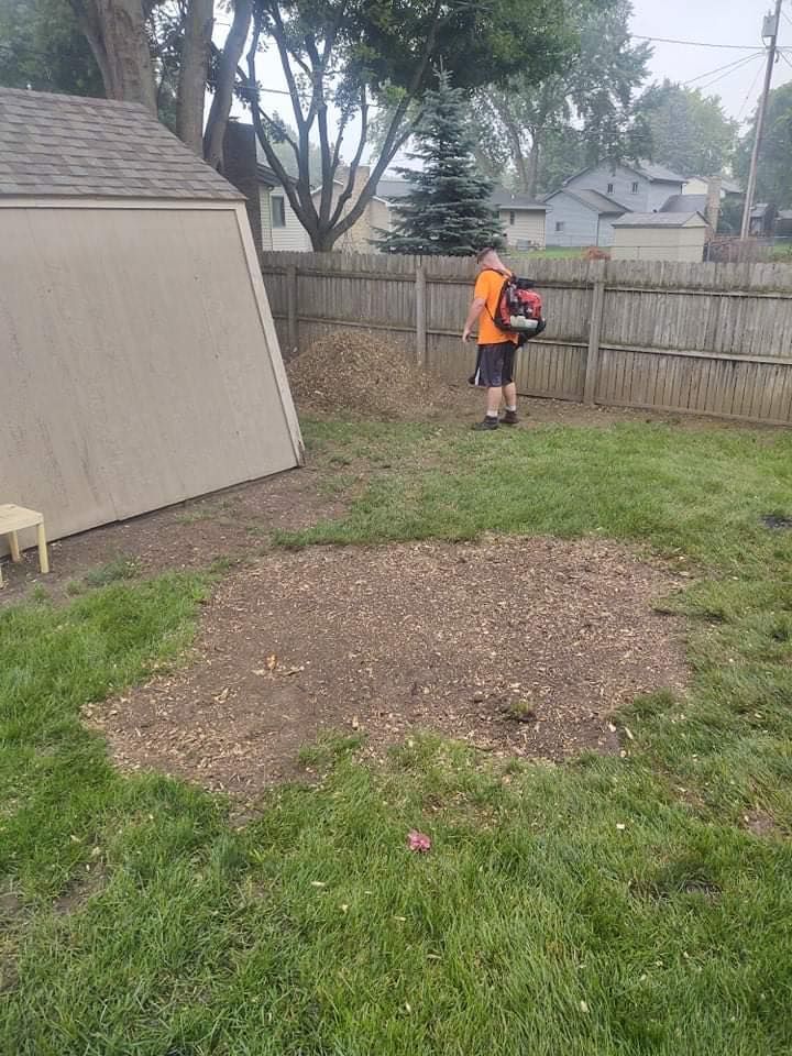A person in orange uses a leaf blower in a backyard with a shed, wooden fence, and mulch.