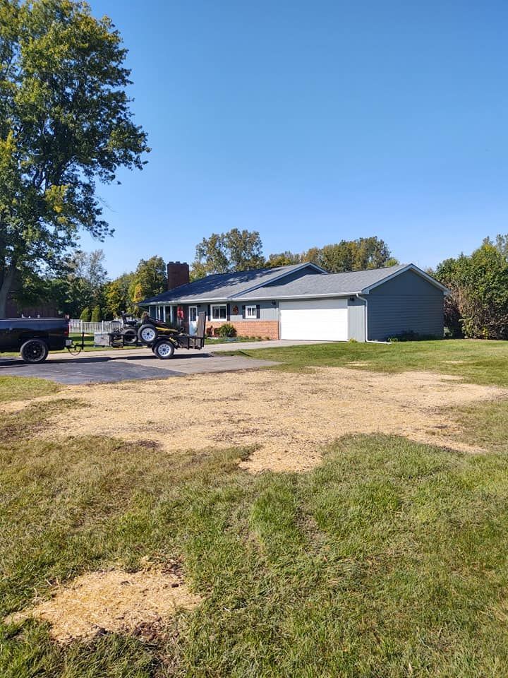A gray-sided house with a white garage door, vehicles in the driveway, and a grassy yard under a blue sky.