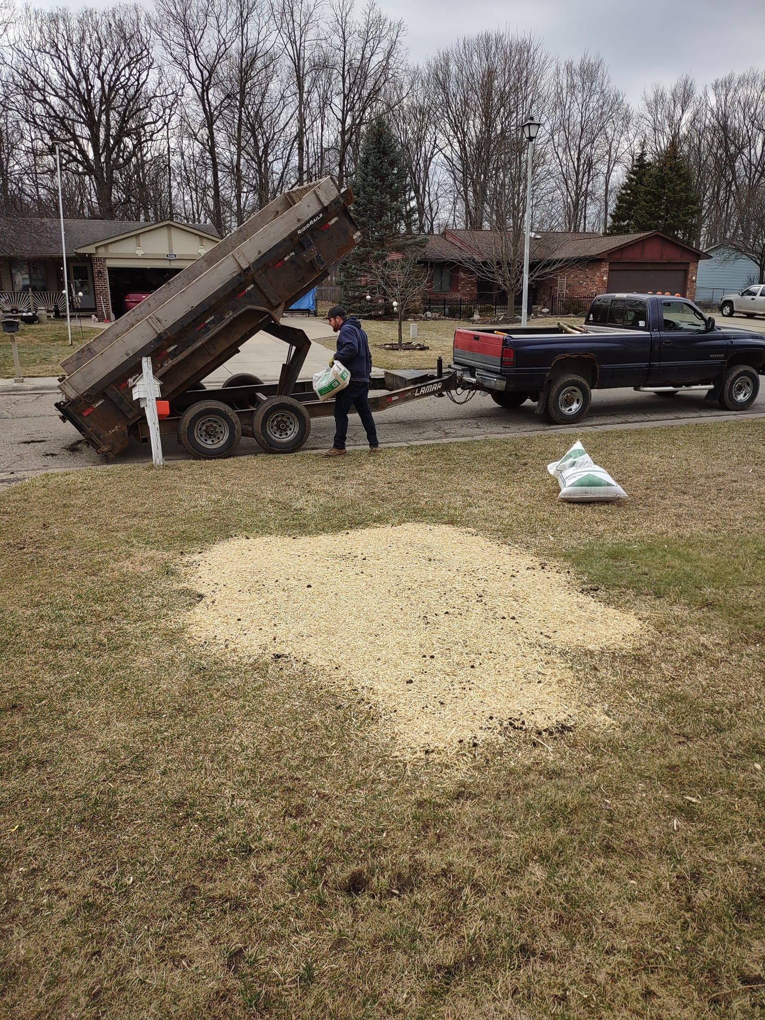 Person unloading gravel from a dump trailer onto a yard, pulled by a pickup truck. Bags of material nearby.