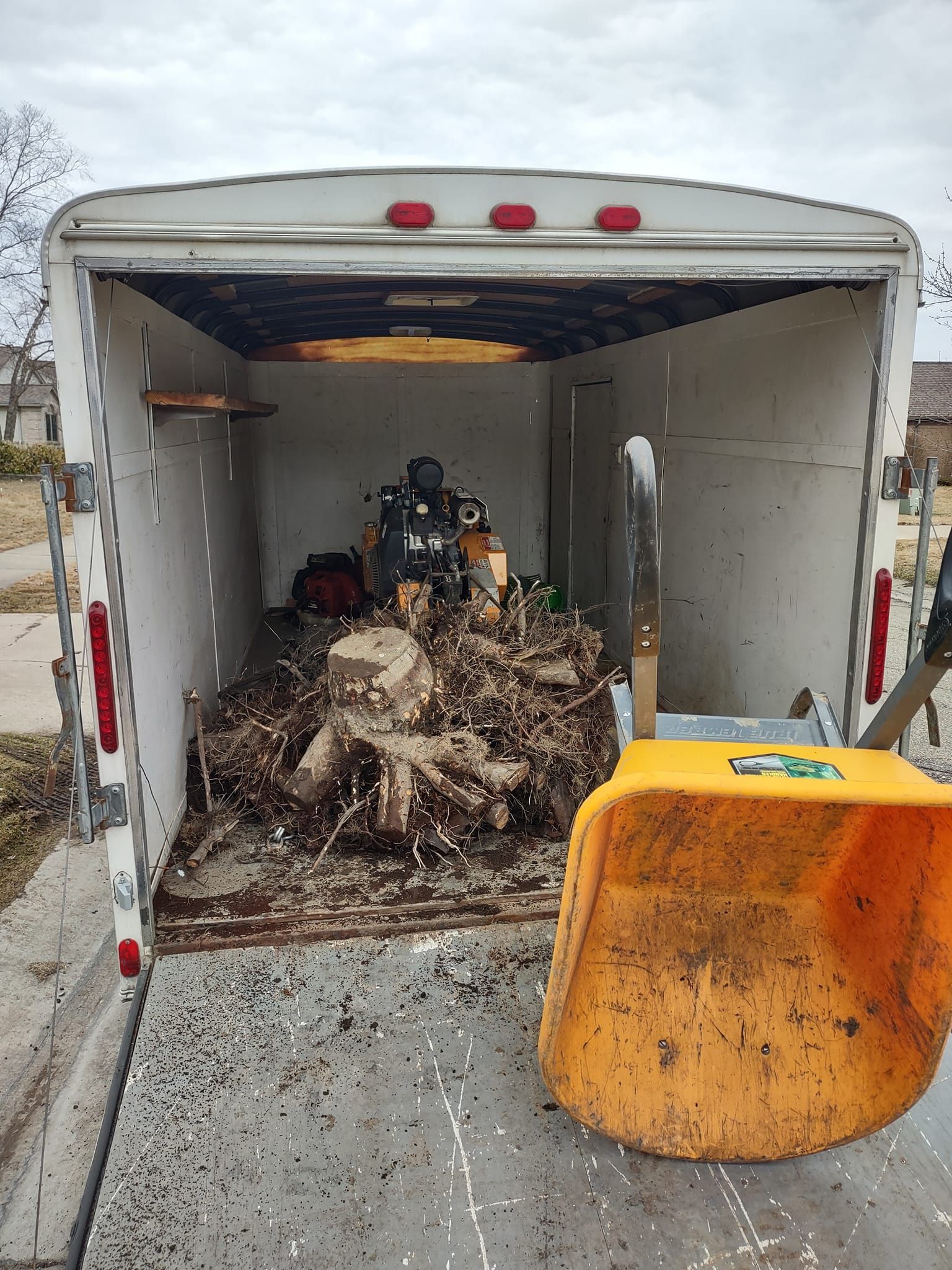 A trailer loaded with wood chips and a stump grinder. An open door and a yellow bucket are visible.