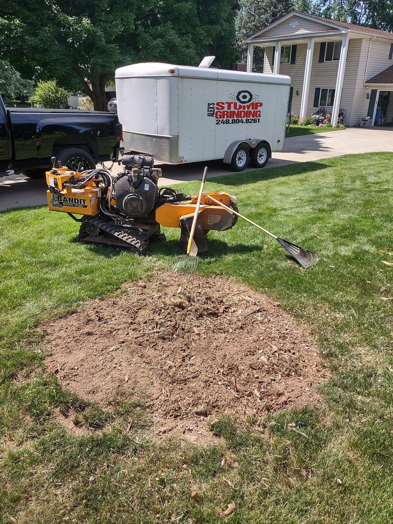 Stump grinder in yard next to a pile of wood chips, with a trailer and truck in the background.