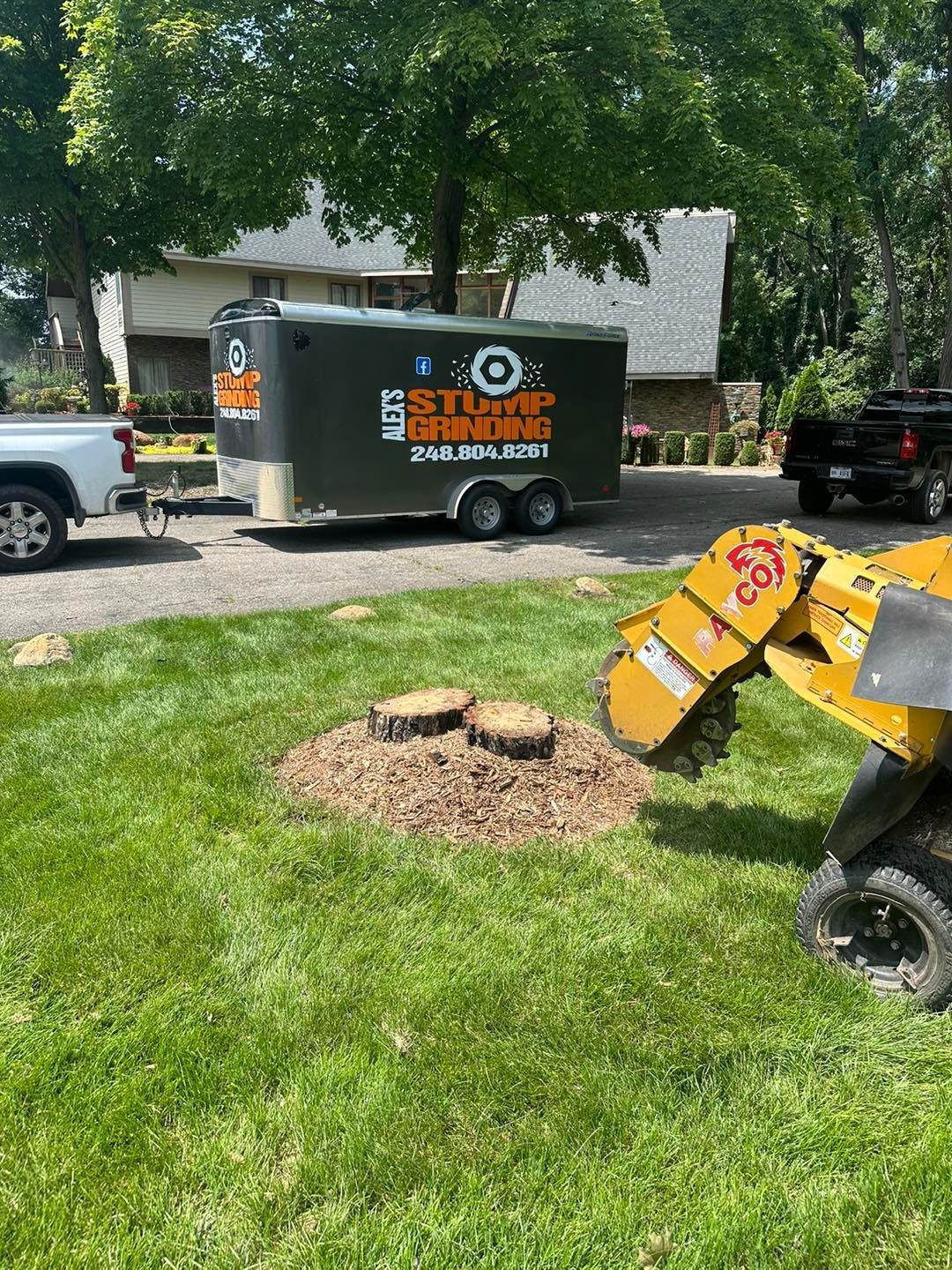 A stump grinder at work on a lawn with a trailer in the background; a truck is also parked nearby.