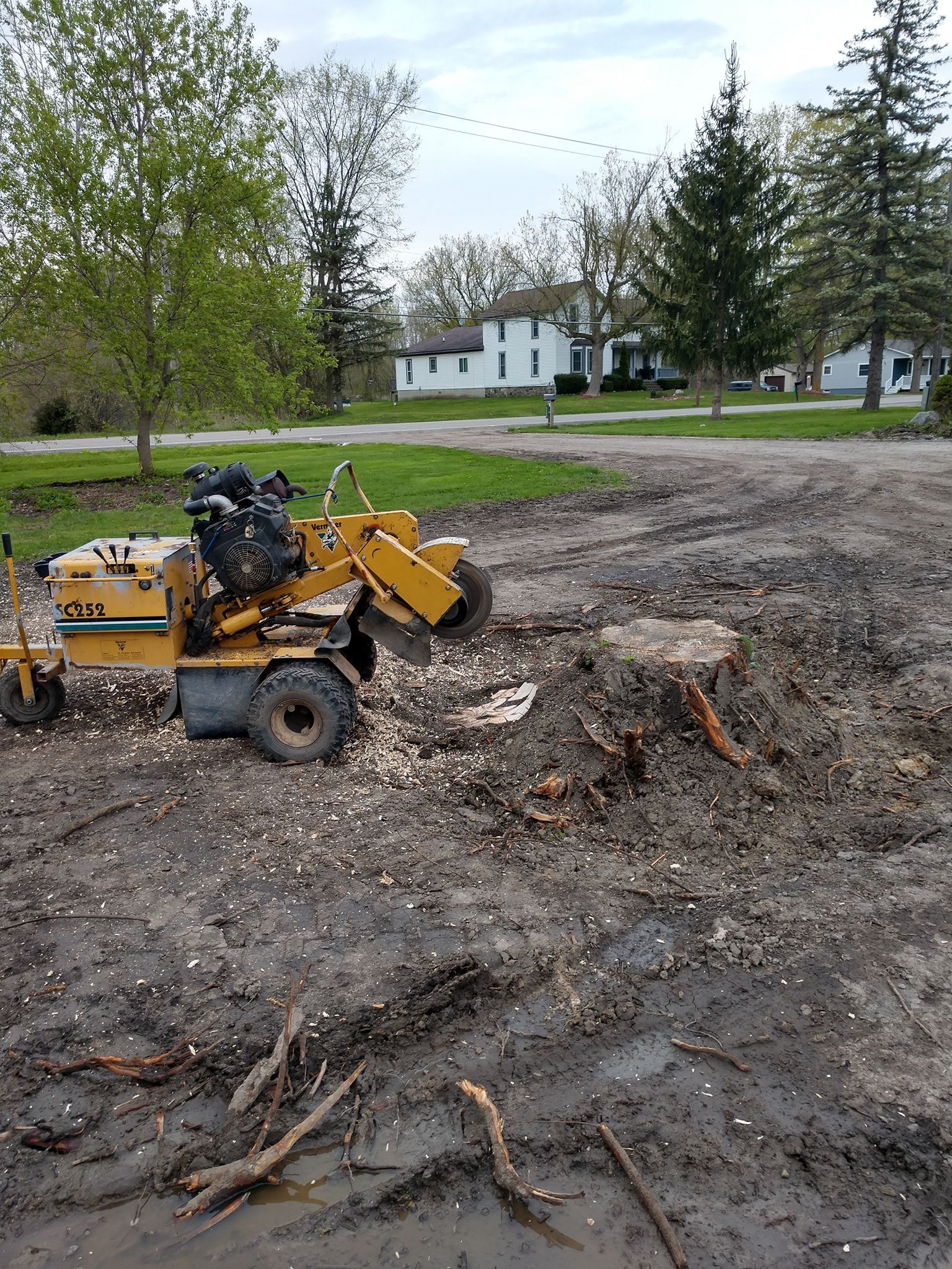 A stump grinder grinding a tree stump in a muddy yard, near a house.