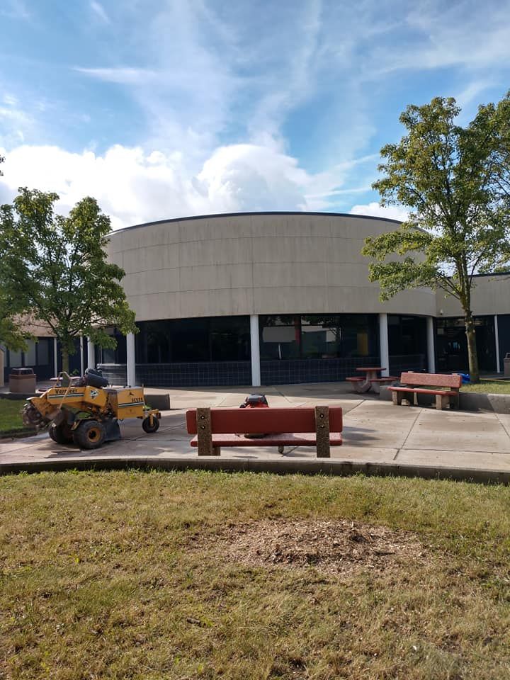 Exterior view of a circular, modern building with a lawnmower, benches, and trees under a cloudy sky.