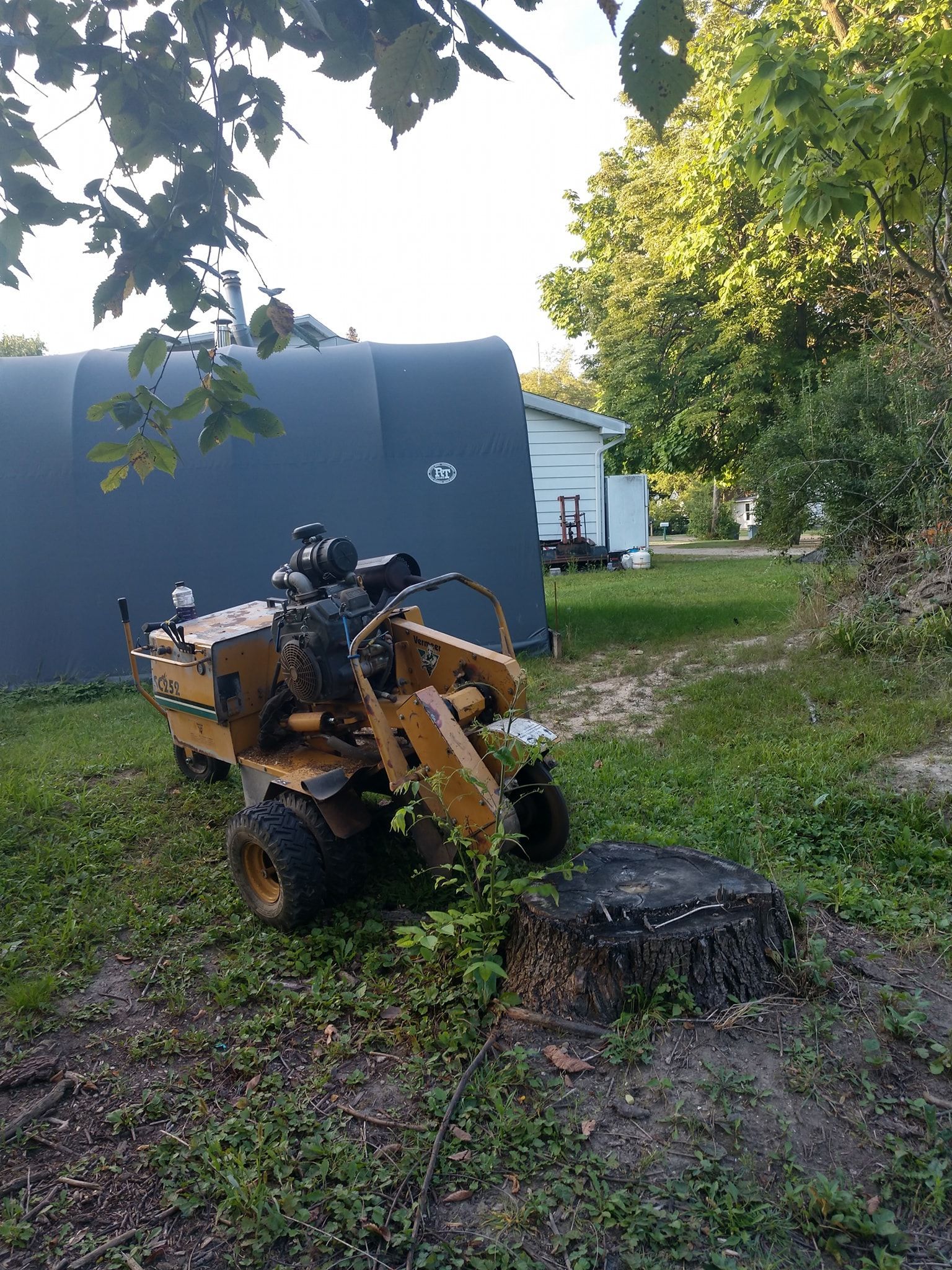 A yellow stump grinder next to a tree stump, with a dark structure and trees in the background.
