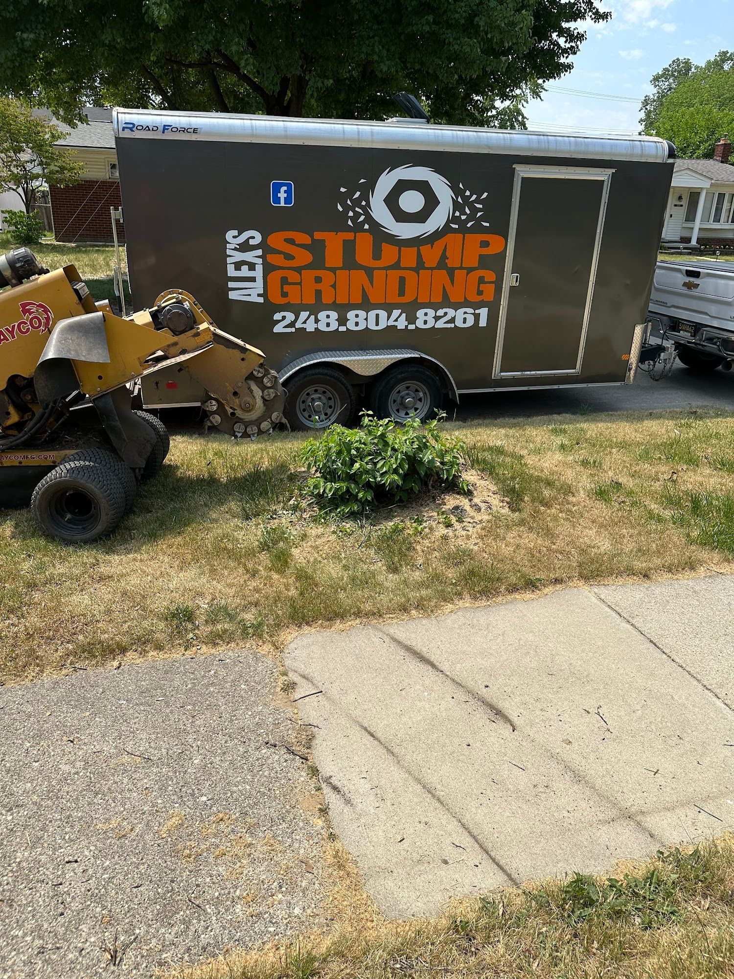 A stump grinding service trailer and equipment parked on a lawn next to a sidewalk.