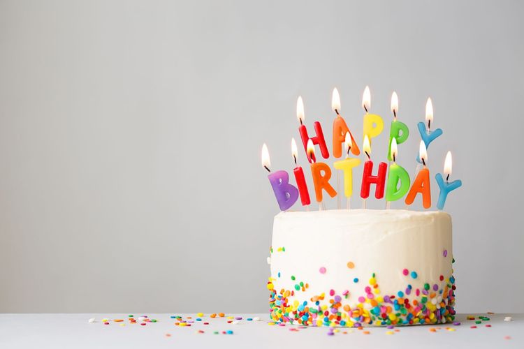 Una torta di compleanno ricoperta di glassa bianca e zuccherini colorati, con sopra delle candeline multicolori accese con la scritta "HAPPY BIRTHDAY".