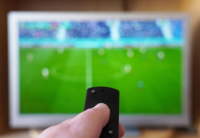 Una mano impugna un telecomando nero, puntandolo verso uno schermo televisivo che mostra un'immagine sfocata di una partita di calcio su un campo.