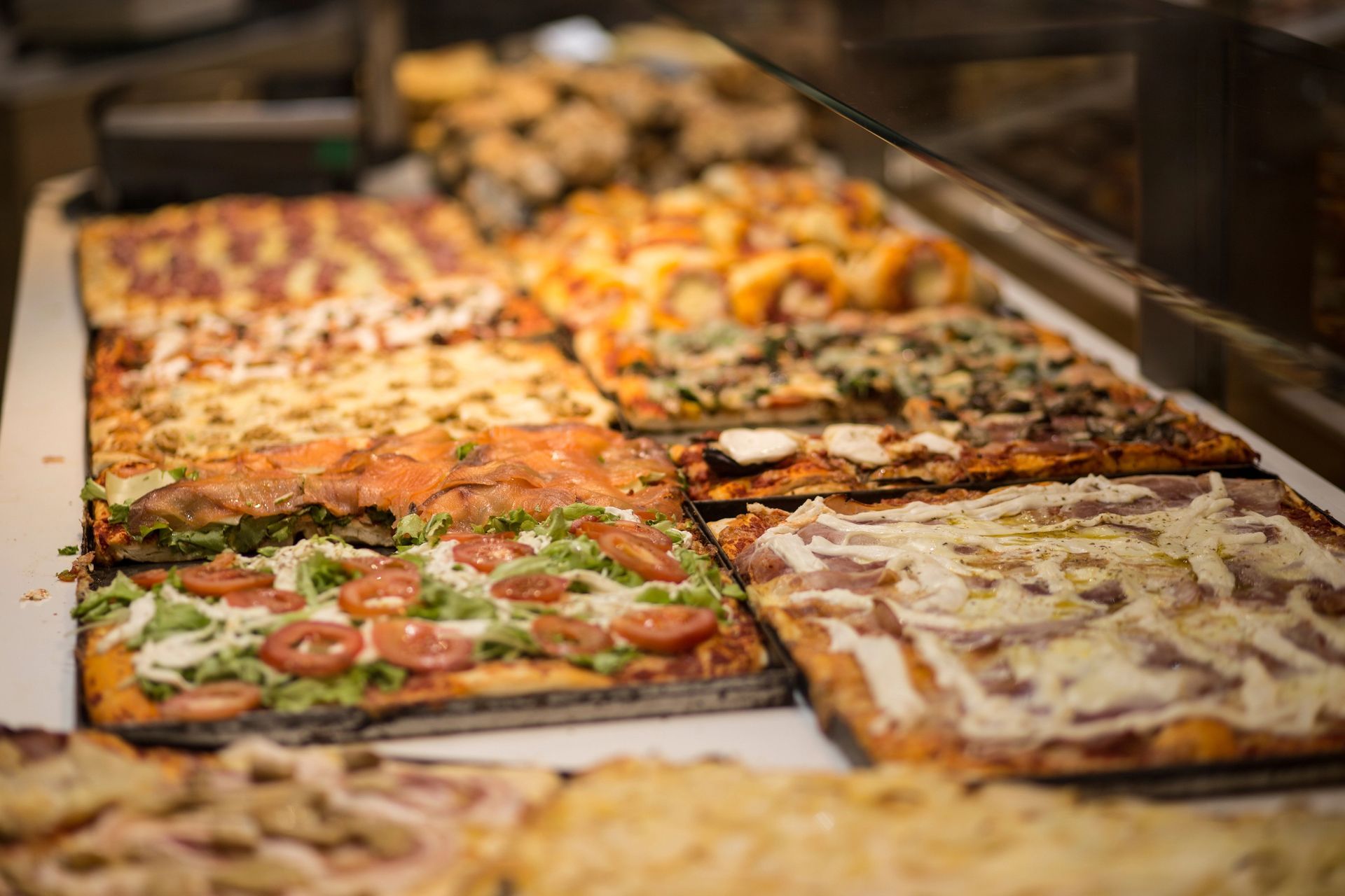 Una vetrina piena di file di pizze quadrate con vari condimenti come pomodoro, verdure e salumi.