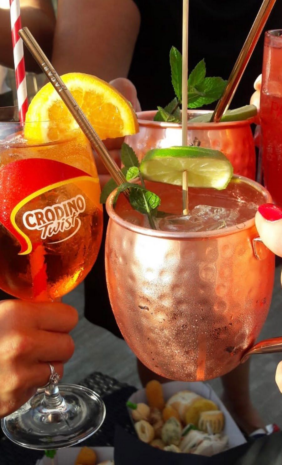 Un primo piano di mani che reggono un cocktail Crodino e una tazza di rame per Moscow Mule, con degli stuzzichini su un tavolo lì vicino.