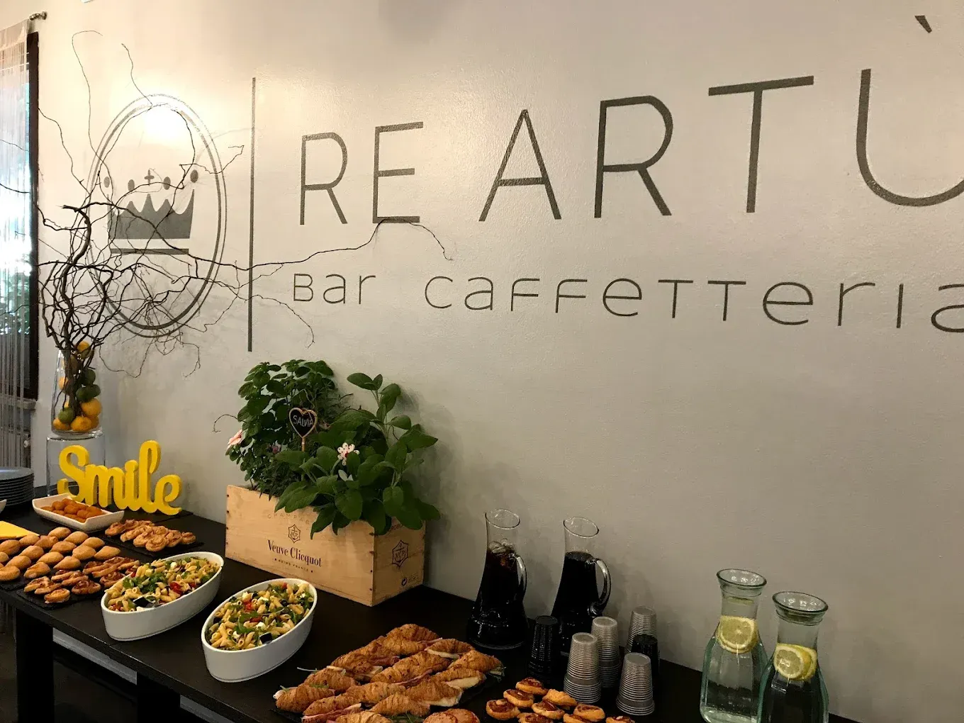 Un tavolo a buffet con antipasti e bevande presso il bar-caffetteria Re Artù, con il nome del locale sulla parete sovrastante.
