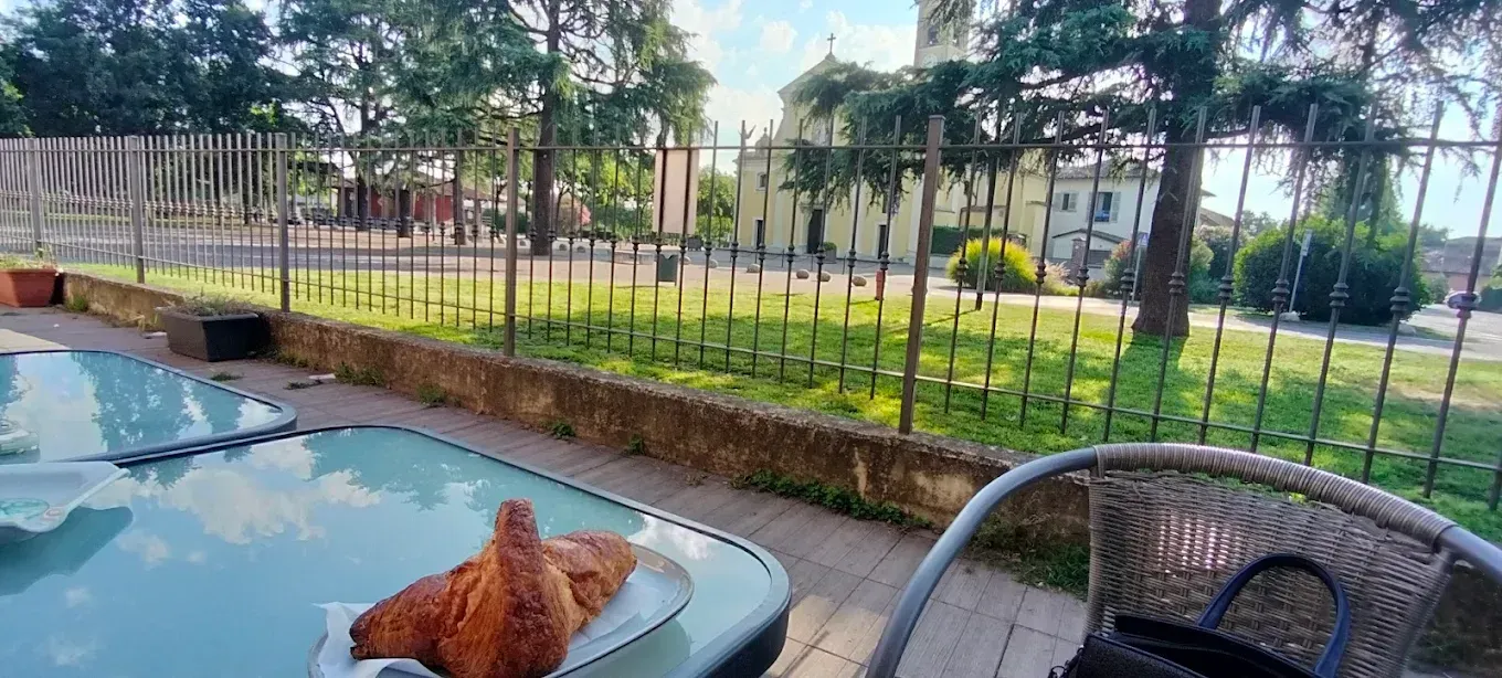 Un croissant su un tavolino di vetro in un caffè all'aperto, con vista su una chiesa e sugli alberi al di là di un prato verde.