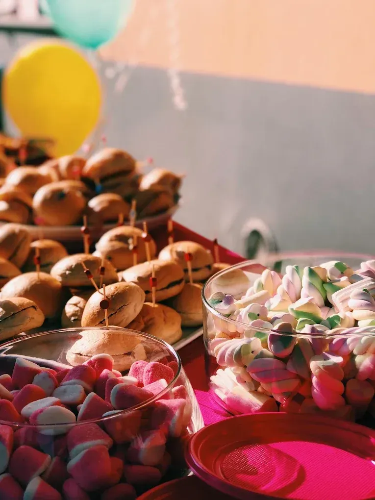 Un buffet per la festa con vassoi di mini-hamburger e ciotole di vetro piene di marshmallow colorati sotto palloncini gialli.