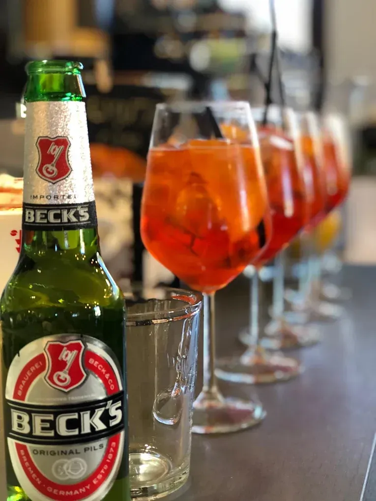 Una bottiglia di birra Beck's accanto a un bicchiere di Aperol Spritz all'arancia, con altri cocktail allineati sullo sfondo.