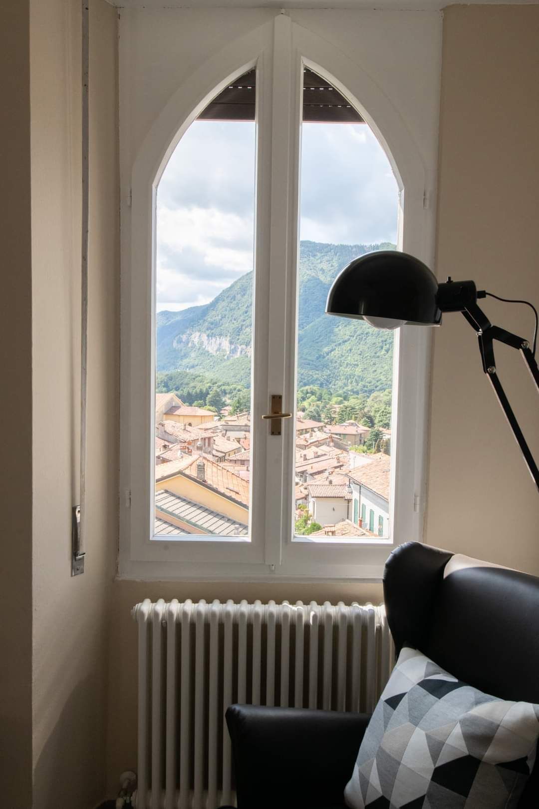 Gothic-style window with a view of a town nestled among mountains. Black lamp and a radiator are nearby.