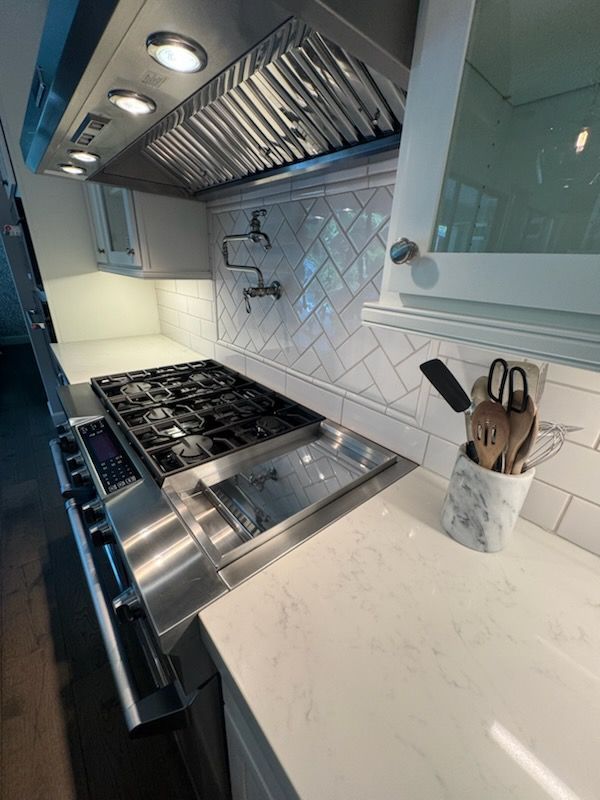Stainless steel kitchen with stovetop, range hood, white countertops and backsplash.