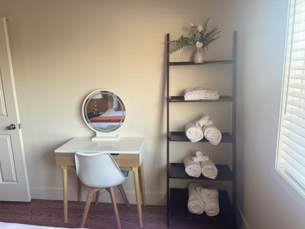 A white desk with a mirror and chair next to a ladder shelf with towels, and a window.